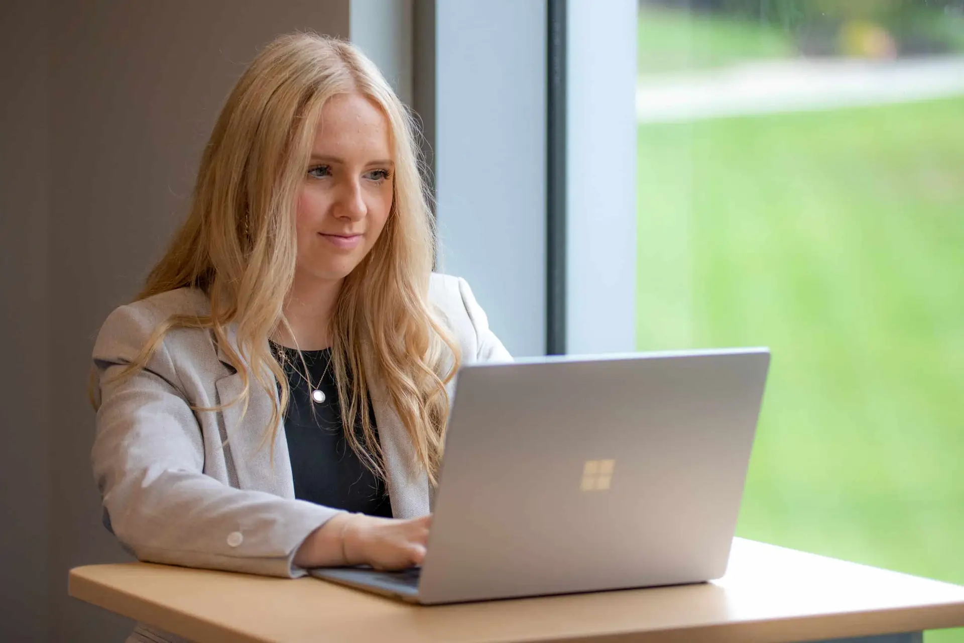 photo of a female student seating next to a window, using her laptop computer